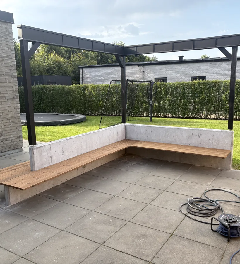Færdig L-formet betonmur med bænk i træ under en sort pergola i haven